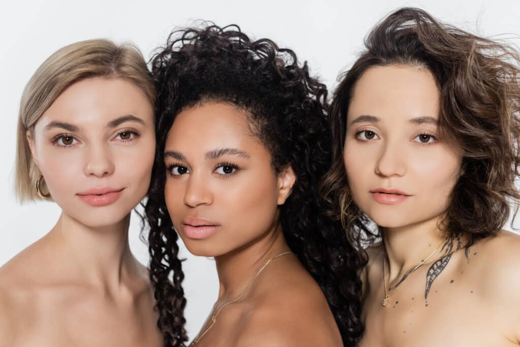 Young multiethnic women with naked shoulders looking at camera isolated on grey